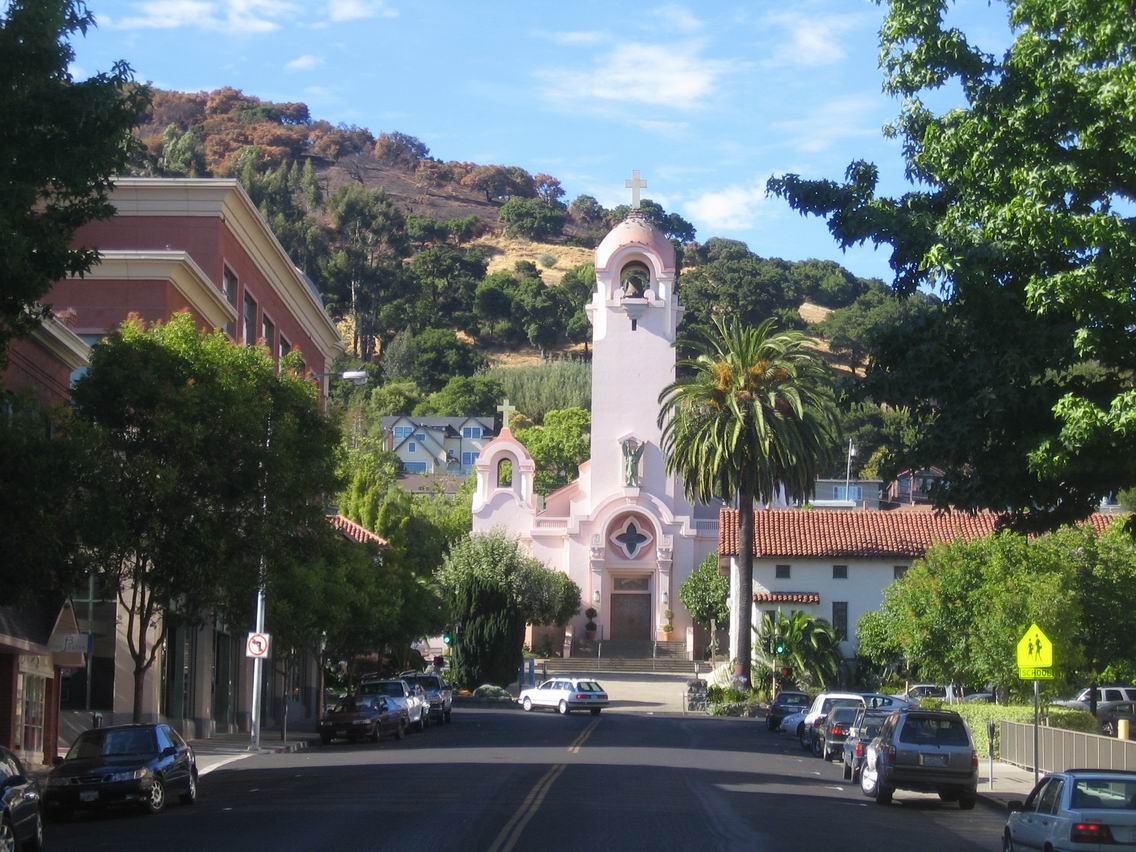 Mission San Rafael Arcángel with pink façade, bell tower, palm trees, and hillside backdrop on a sunny day in downtown San Rafael, CA.